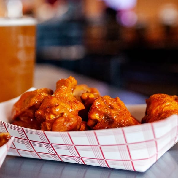 Wings to Go's saucy chicken wings in a paper tray with a blurred beer in the background.