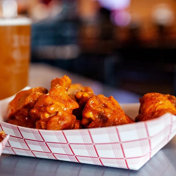Close-up of saucy chicken wings in a red and white checkered paper tray, with a drink in the background.