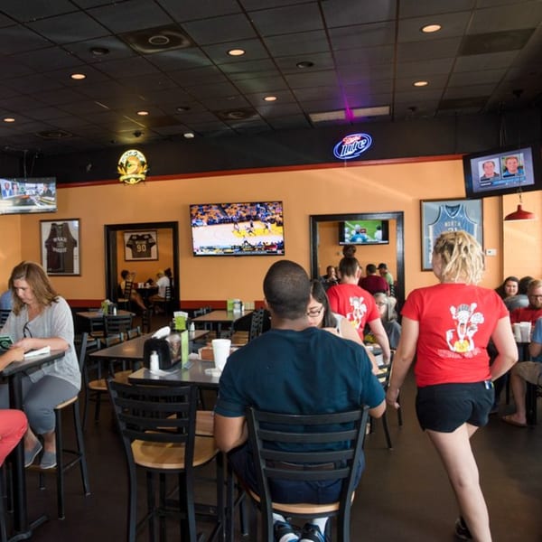 Diners seated at tables in a casual restaurant with multiple TVs showing sports and neon signs.