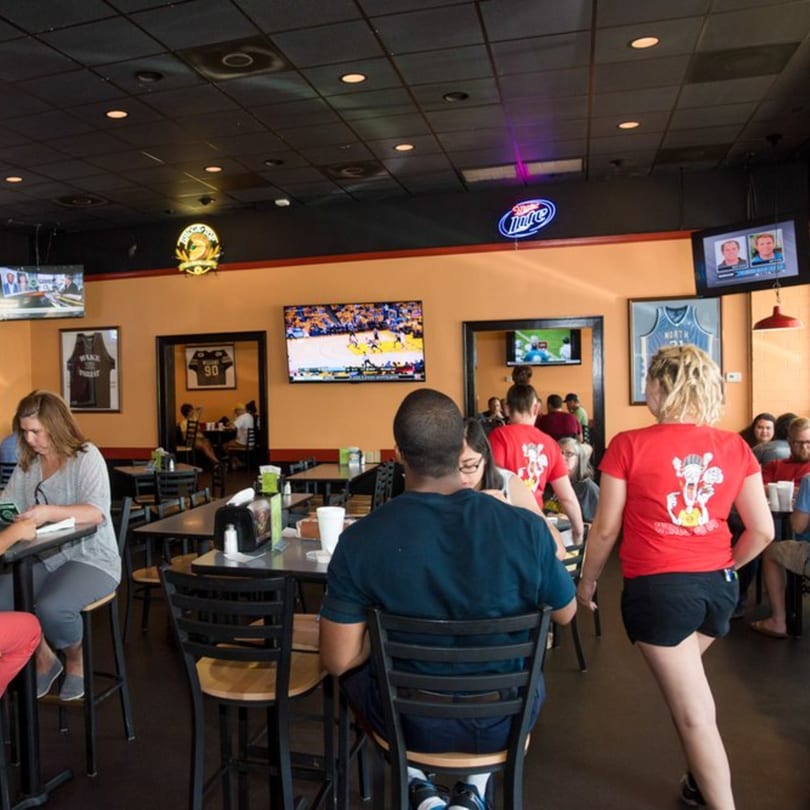 Diners seated at tables in a casual restaurant with multiple TVs showing sports and neon signs.