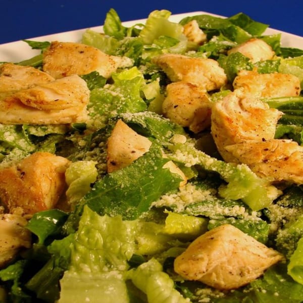 Close-up of a Caesar salad with grilled chicken pieces and grated cheese on a white plate.